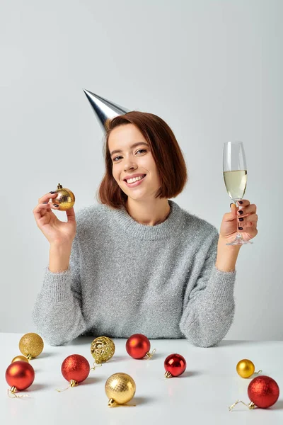 Delighted woman in party cap holding champagne glass and bauble on grey, Merry Christmas concept — Stock Photo
