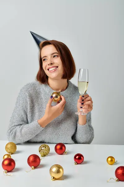 Mujer complacida en la tapa del partido sosteniendo copa de champán y bauble en gris, Feliz concepto de Navidad - foto de stock