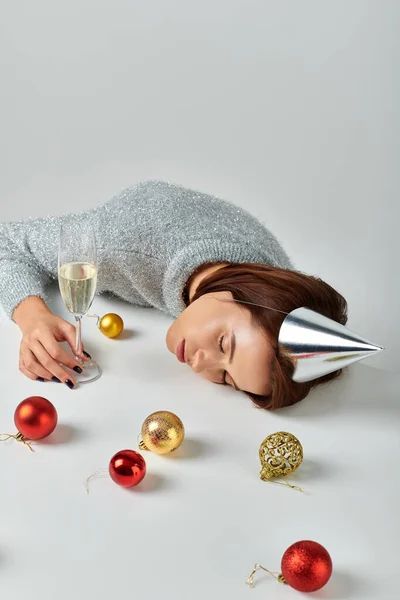 Woman in party cap sleeping on table near champagne glass and Christmas baubles on grey backdrop — Stock Photo