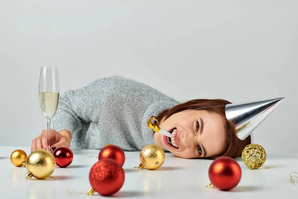 Woman in party cap lying on table near champagne glass and Christmas baubles, blowing party horn — Stock Photo