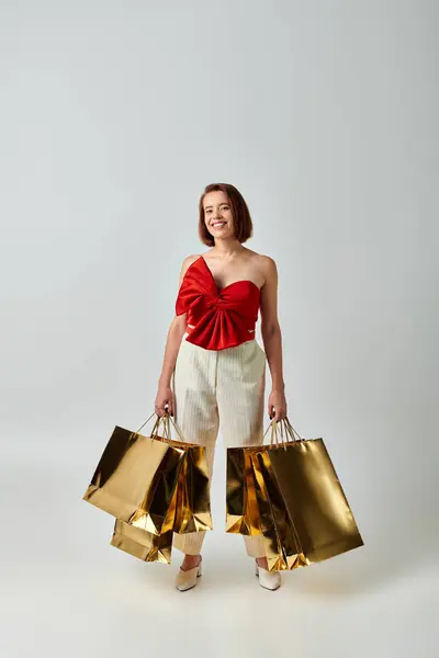 Shopping natalizio, donna felice in top rosso alla moda senza spalline con fiocco che tiene borse della spesa sul grigio — Foto stock