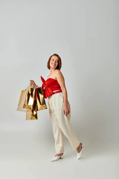 Compras de Año Nuevo, mujer joven feliz con un atuendo elegante sosteniendo bolsas de compras sobre fondo gris - foto de stock
