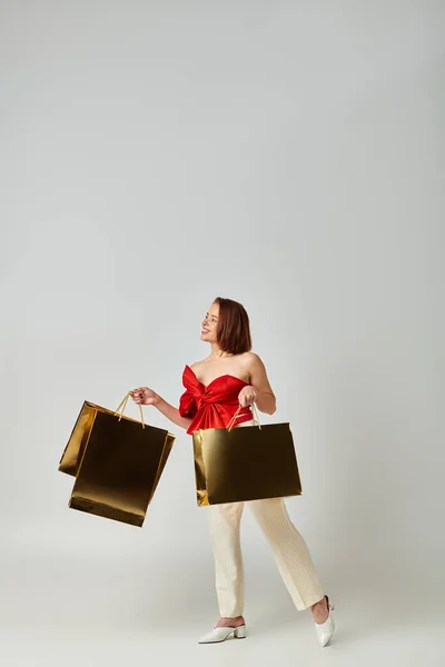 Compras de Navidad, mujer joven positiva en traje de moda sosteniendo bolsas de compras sobre fondo gris - foto de stock