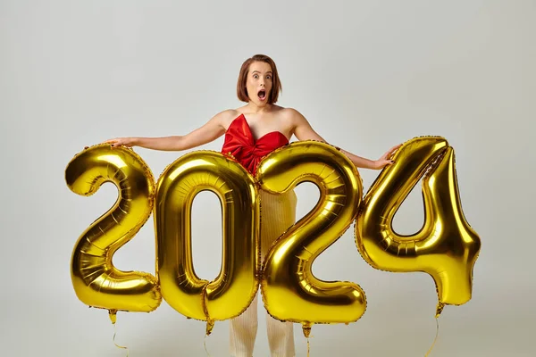 Happy New Year, shocked young woman in trendy attire holding balloons with 2024 numbers on grey — Stock Photo