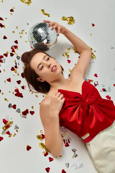 Festa de Ano Novo, jovem morena com bola de discoteca deitada no chão perto de confetes em pano de fundo cinza — Fotografia de Stock