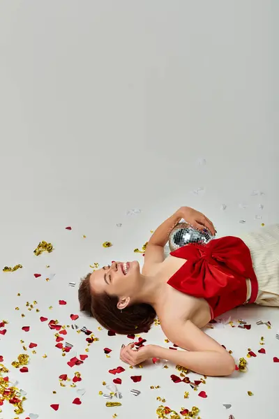 Festa de Ano Novo, jovem excitada com bola de discoteca deitada no chão perto de confete em pano de fundo cinza — Fotografia de Stock