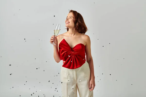 Feliz Ano Novo, mulher alegre em vestido de festa segurando copo de champanhe perto de confete em cinza — Fotografia de Stock