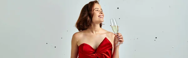 New Year banner, cheerful woman in party attire holding glass of champagne near confetti on grey — Stock Photo