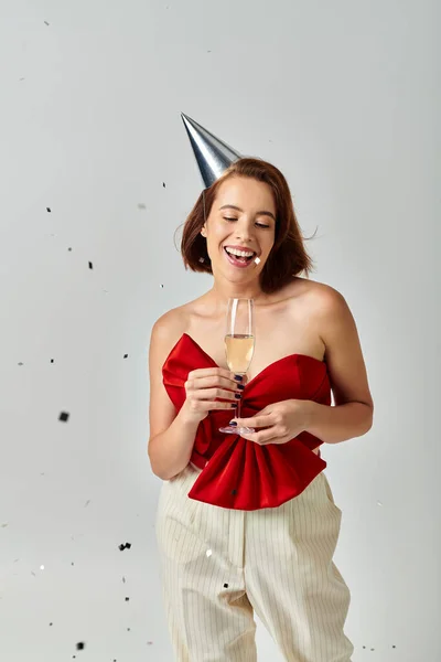 Happy New Year, positive young woman in party cap holding glass of champagne near confetti on grey — Stock Photo
