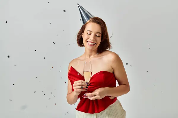 Merry Christmas, positive young woman in party cap holding glass of champagne near confetti on grey — Stock Photo