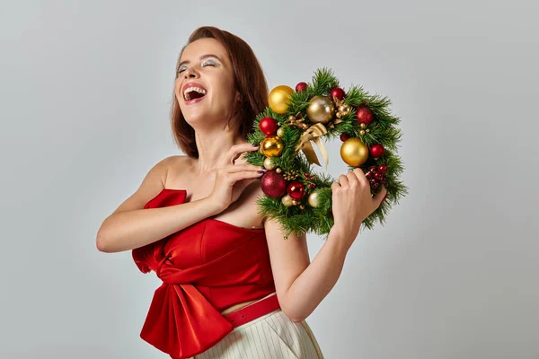 Festive season, inspired woman with shimmery makeup holding Christmas wreath on grey backdrop — Stock Photo