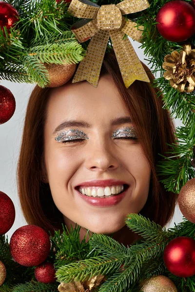 Close up view of happy woman with shimmery makeup smiling inside frame of decorated Christmas wreath — Stock Photo