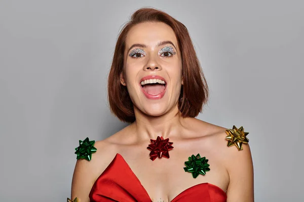 Feliz Navidad, mujer excitada en traje rojo con arcos por todo su cuerpo riendo sobre fondo gris - foto de stock