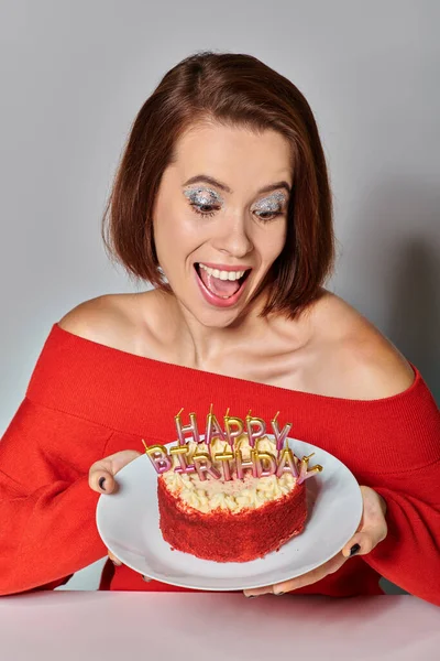 Thrilled woman in red attire looking at bento cake with Happy Birthday candles on grey background — Stock Photo