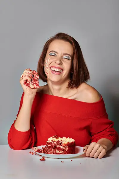 Chica de cumpleaños positivo con los ojos cerrados comiendo delicioso pedazo de pastel de terciopelo rojo sobre fondo gris - foto de stock