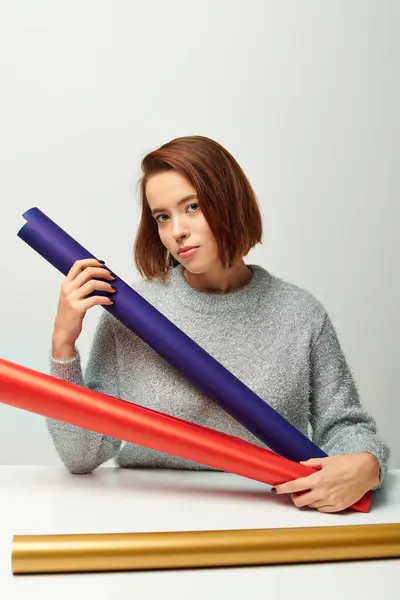 Pretty woman in sweater choosing between different colors of Christmas gift paper on grey backdrop — Stock Photo