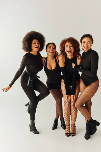 Jóvenes mujeres afroamericanas alegres en trajes sexy negro posando felizmente en movimiento, concepto de moda - foto de stock