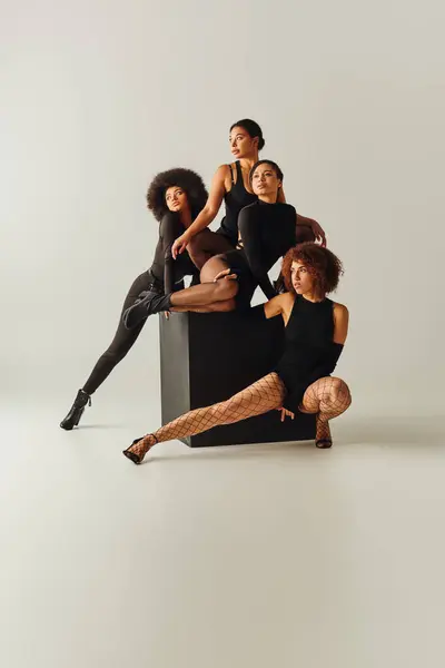 Atractivo afroamericano mujeres en sexy bodysuits posando juntos en negro cubo, concepto de moda - foto de stock