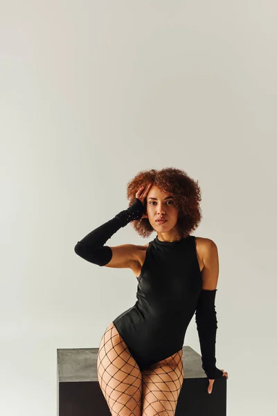 Atractiva mujer afroamericana en medias de rejilla y traje de cuerpo sexy posando cerca de cubo negro, moda - foto de stock
