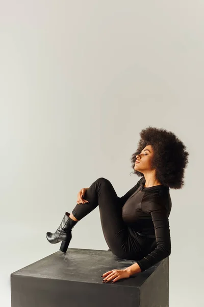 Mulher americana africana atraente em preto sedutor bodysuit sentado no cubo preto, conceito de moda — Fotografia de Stock
