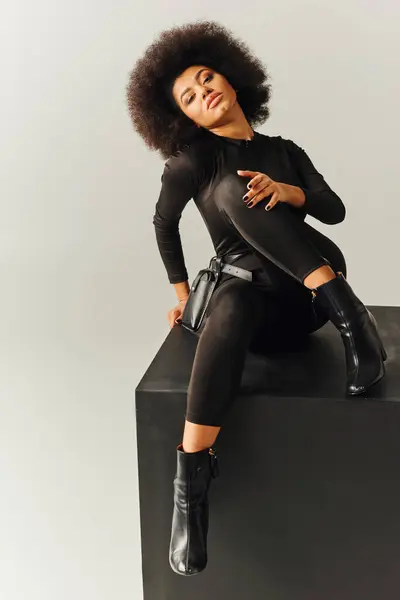 Attraktive afrikanisch-amerikanische Frau im schwarzen, verführerischen Body auf schwarzem Würfel sitzend, Modekonzept — Stockfoto