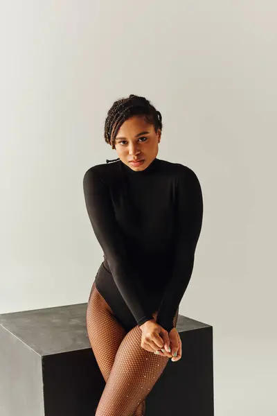 Sexy joven afroamericana mujer en seductor traje negro sentado en cubo negro, concepto de moda - foto de stock