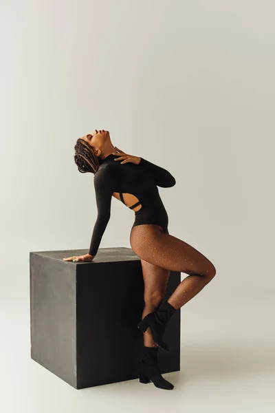 Alluring african american woman in sexy tights and bodysuit posing next to black cube, fashion — Stock Photo