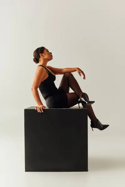 Schöne afrikanisch-amerikanische Frau in verführerisch stilvollen Body sitzt auf schwarzen Würfel, Modekonzept — Stockfoto