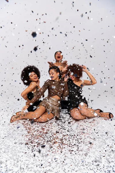 Mujeres afroamericanas atractivas elegantes que festejan bajo la lluvia del confeti que se sienta en piso, moda - foto de stock