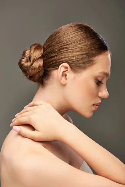Vista lateral da jovem com coque de cabelo e pele perfeita posando em fundo cinza, encantador — Fotografia de Stock