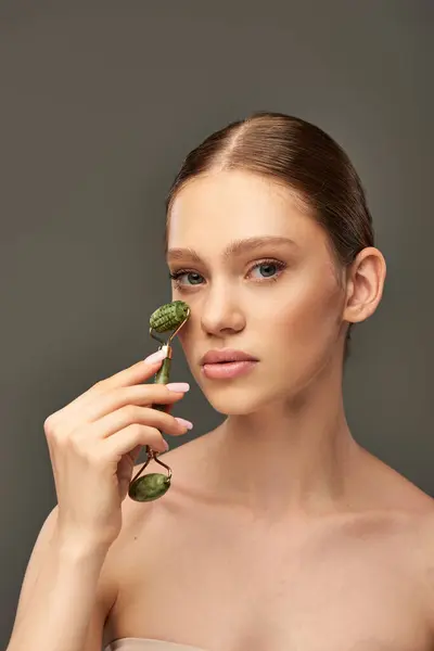 Giovane donna con pelle perfetta massaggio viso con rullo di giada su sfondo grigio, ringiovanimento della pelle — Foto stock
