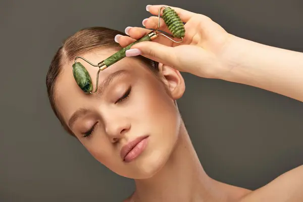 Jeune femme avec peau parfaite massage visage avec rouleau de jade sur fond gris, choyer la peau — Photo de stock