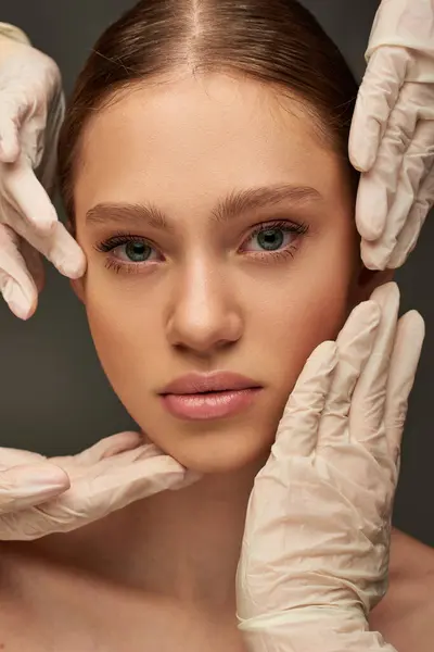 Gros plan d'esthéticiennes en gants médicaux examinant le visage d'une jeune jolie patiente sur fond gris — Photo de stock
