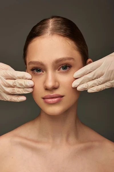 Estheticians in medical gloves touching face of pretty woman on grey background, dermatology concept — Stock Photo