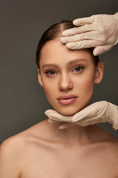 Esthéticienne en gants médicaux touchant le visage d'une jeune femme sur fond gris, concept dermatologique — Photo de stock