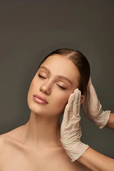 Esthetician in medical gloves touching face of young woman on grey background, facial treatment — Stock Photo
