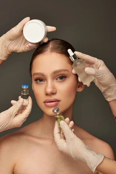 Estheticians in medical gloves holding different beauty treatment products near young woman on grey — Stock Photo