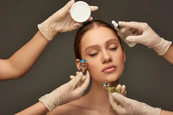 Estheticians in medical gloves holding beauty treatment products near woman on grey backdrop — Stock Photo