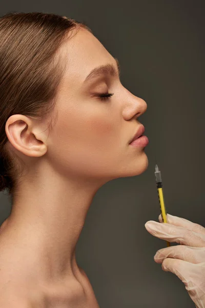 Esthetician in medical glove holding syringe near lips of young woman on grey background, side view — Stock Photo