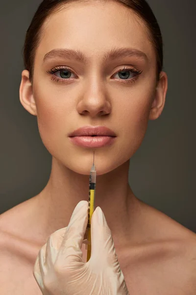 Esthetician in medical glove holding syringe near young woman on grey background, lip filler — Stock Photo