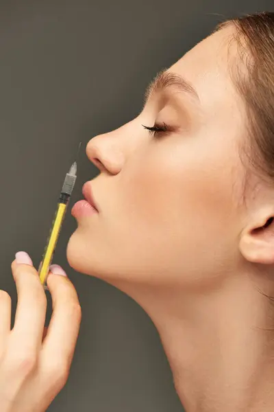 Retrato da jovem mulher segurando seringa perto do rosto em fundo cinza, conceito de realce labial — Fotografia de Stock