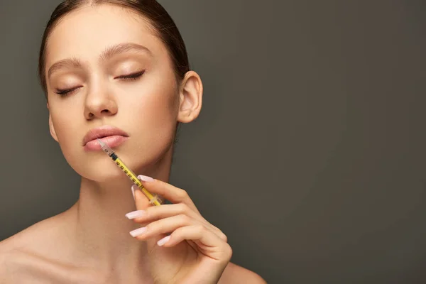 Portrait de jolie jeune femme tenant une seringue près du visage sur fond gris, concept de mise en valeur des lèvres — Photo de stock