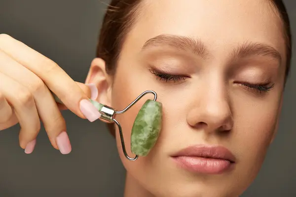 Cerrar mujer joven con los ojos cerrados haciendo masaje facial con rodillo de jade verde sobre fondo gris - foto de stock