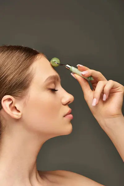 Side view of young woman with closed eyes doing face massage with jade roller on grey background — Stock Photo