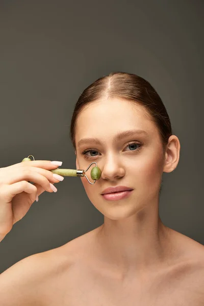 Портрет молодой женщины, делающей массаж носа с зеленым нефритовым роликом на сером фоне, уход за собой — стоковое фото