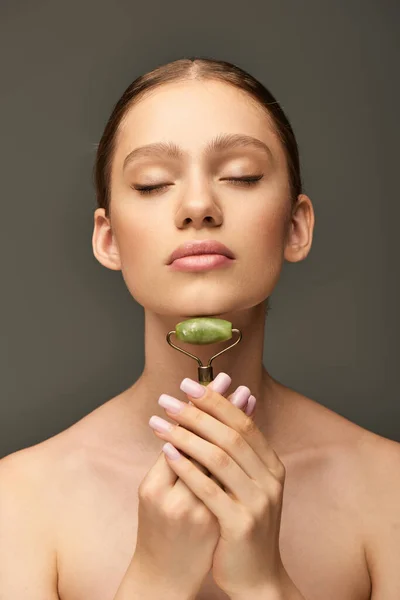 Portrait of young woman doing massage of neck with green jade roller on grey background, self-care — Stock Photo
