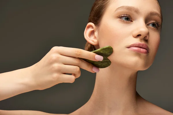 Jeune femme faisant auto-massage avec rouleau de jade vert sur fond gris, soins de la peau et beauté — Photo de stock