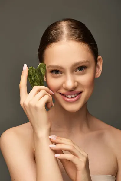Happy young woman smiling and holding jade gua sha stone on grey background, facial contour concept — Stock Photo