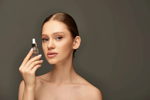 Hübsche Frau mit glühender Haut, Flasche mit Serum auf grauem Hintergrund, Hydratation und Hautpflege — Stockfoto