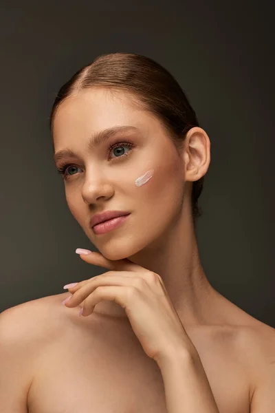 Pretty young woman with cream on face and bare shoulders posing on grey background, look away — Stock Photo